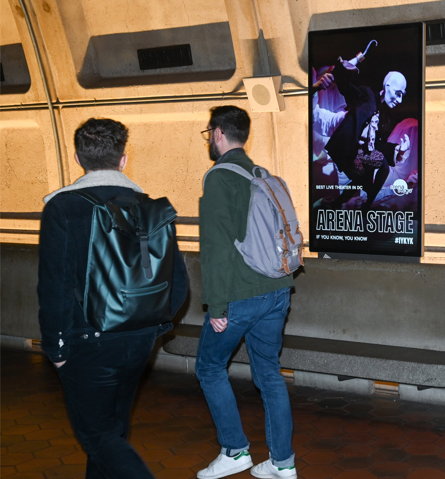 a photo taken on a Metro Platform in Washington, DC, with two people walking past a digital billboard that shows an Arena Stage digital ad.